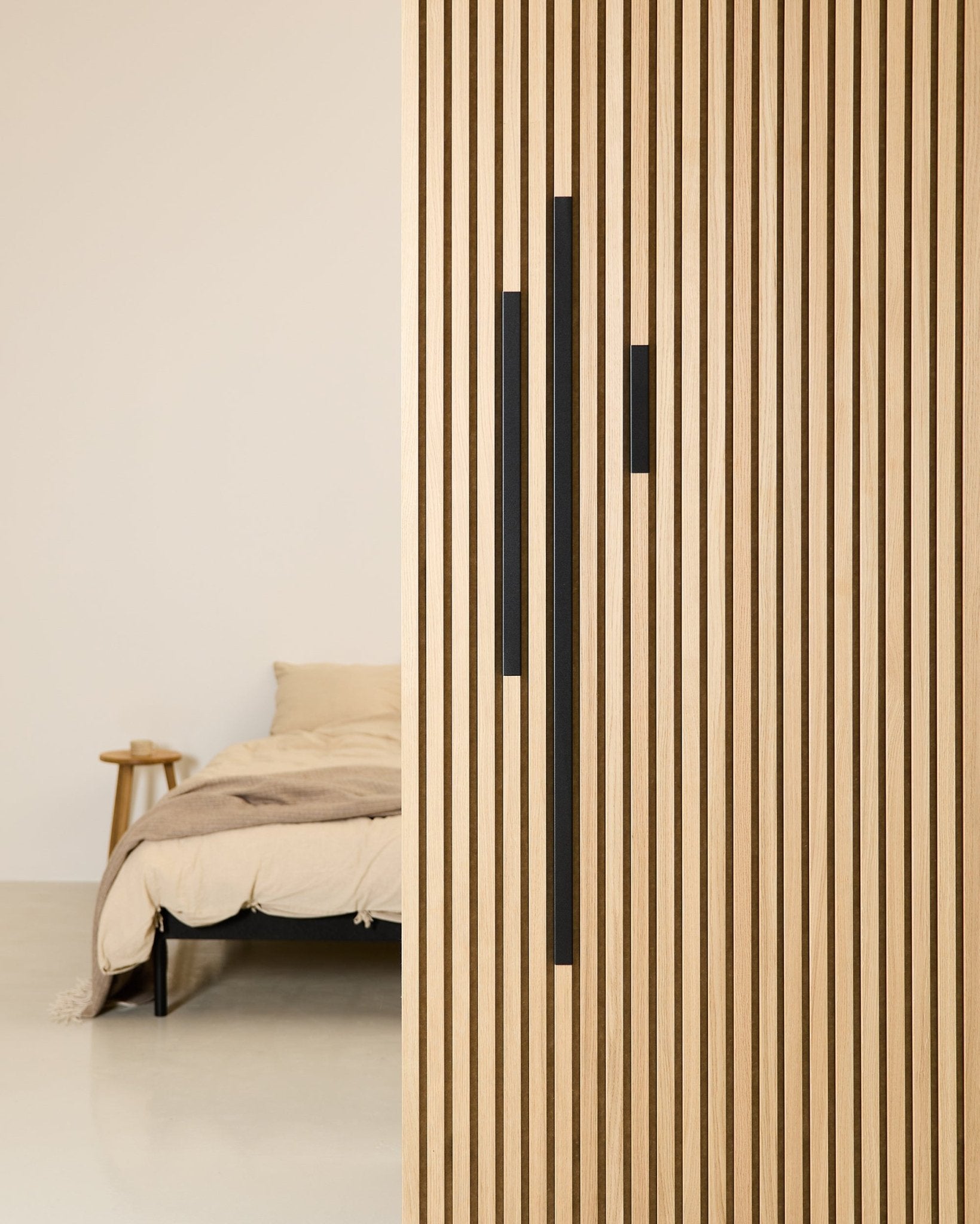 Camera da letto minimalista con letto e coperta beige, parzialmente nascosti da un divisorio WoodUpp Akupanel da 60 cm in alluminio Black della serie Liva Cover.